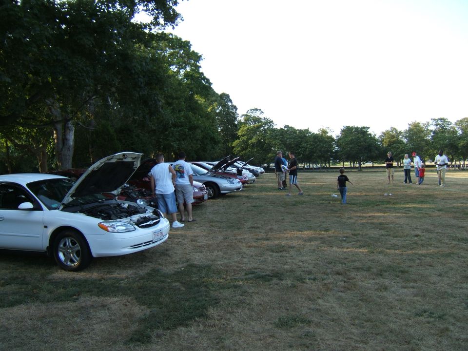 White sedan with hood up, parked in a line with other cars. People stand around and talk