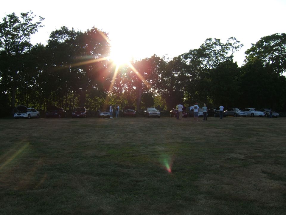 Several cars parked in a grassy field with trees and people gathered