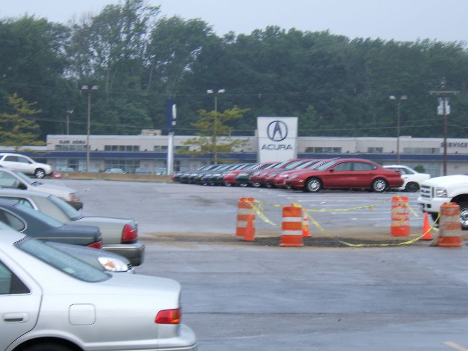 Parking lot in front of an Acura dealership with several cars parked in rows and a white truck