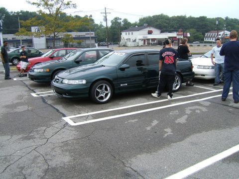 1998 Ford Taurus - Several sedans are parked in a lot, with people standing around them.