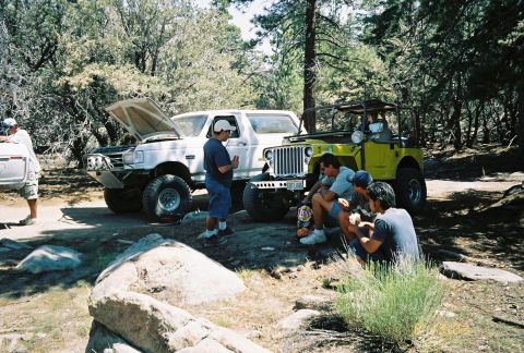 1990 Ford Bronco - Off-roaders take a break by a white lifted truck with its hood up and a yellow flatfender Jeep in a pine clearing.
