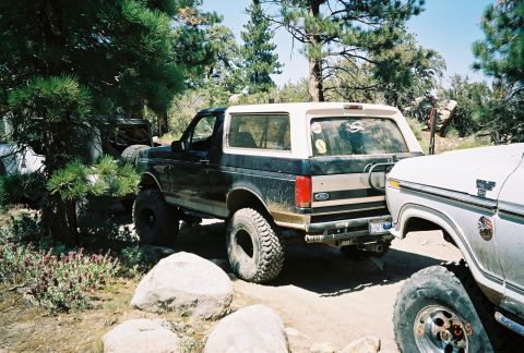 1990 Ford Bronco - dark lifted Ford Bronco on oversized tires is parked next to a white pickup on a rocky pine-lined trail.
