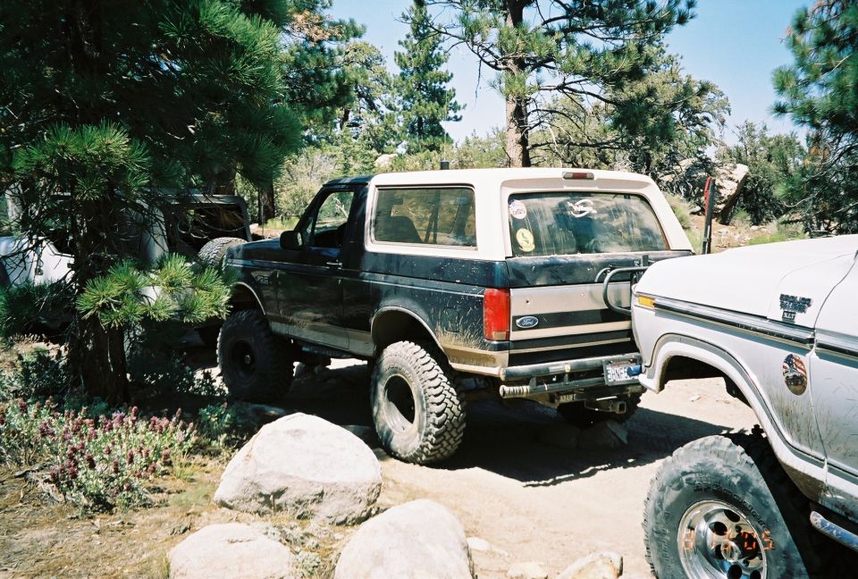 Dark lifted Ford Bronco on big tires parked beside a white pickup on a rocky pine trail