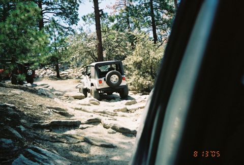 1990 Ford Bronco - white Jeep Wrangler with a rear spare climbs a rocky trail, framed by another vehicle's door pillar.