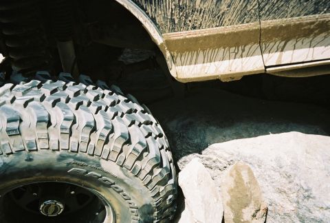 1990 Ford Bronco - close-up of a mud-splattered BFGoodrich off-road tire on a lifted vehicle wedged beside a large rock.