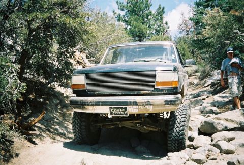 1990 Ford Bronco - dusty lifted Ford truck with a billet grille and 'Trail This!' plate flexes over rocks as a man walks nearby.