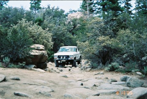 1990 Ford Bronco - white lifted SUV on oversized tires sits on a dirt trail surrounded by high-desert brush and boulders.