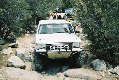 1990 Ford Bronco - white lifted SUV fitted with a tube bumper and three driving lights crawls over rocks on a brushy trail.