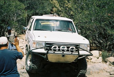 1990 Ford Bronco - man spots a white lifted SUV with a tube bumper and driving lights as it crawls over rocks on the trail.