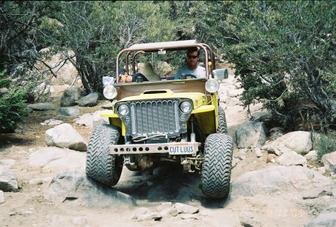 1990 Ford Bronco - yellow flatfender Jeep with a roll cage and 'CUT LUUS' license plate crawls over rocks on a wooded trail.