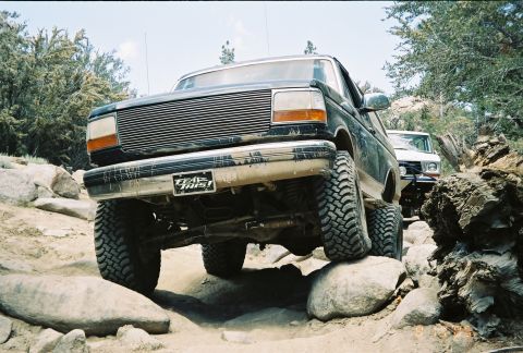 1990 Ford Bronco - From a low angle, a dark lifted Ford truck with a billet grille and 'Trail This!' plate climbs over large boulders.