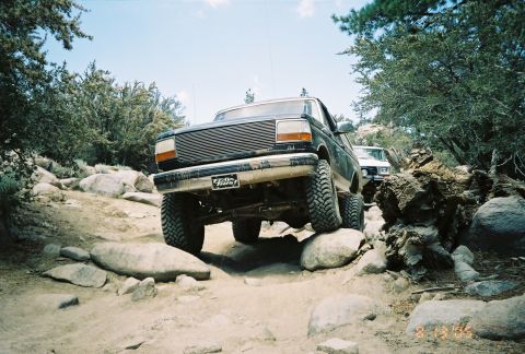 1990 Ford Bronco - dark lifted Ford truck with a billet grille and 'Trail This!' plate flexes a wheel high while crawling over rocks.