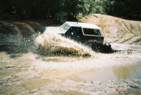1990 Ford Bronco - two-tone SUV charges through a muddy water hole, throwing up a tall sheet of brown spray.