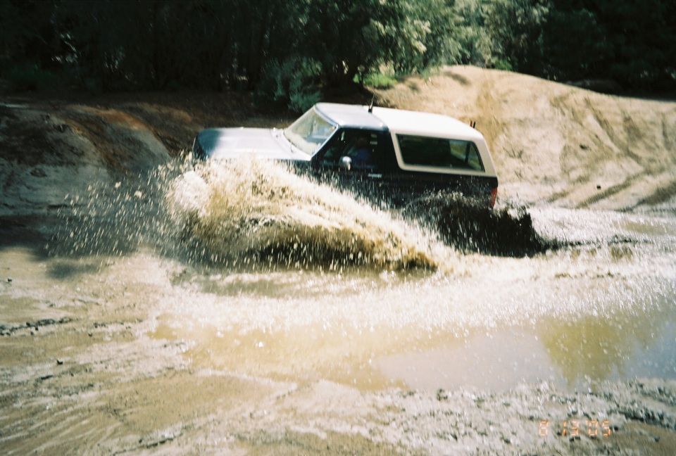 Two-tone SUV blasting through a muddy water pit sending up a large spray