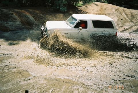 1990 Ford Bronco - white SUV with a red interior powers through a mud pit, flinging up a wide spray of mud and debris.