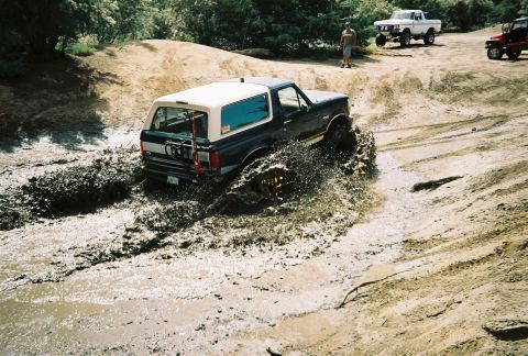 1990 Ford Bronco - dark Ford Bronco with a white top churns through muddy water on a dirt course as others watch from the bank.