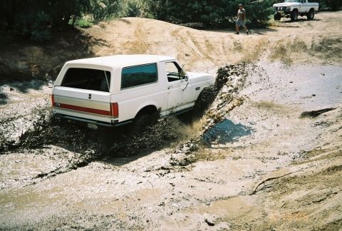 1990 Ford Bronco - Seen from above, a white SUV with a red-striped top wades through a muddy water crossing on a dirt trail.