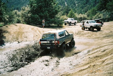 1990 Ford Bronco - dark Ford Bronco with a tan top crosses a shallow muddy stream while several 4x4s wait on the far bank.