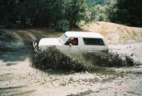 1990 Ford Bronco - white SUV with a red interior plows through a mud pit, sending a broad fan of mud across the dirt course.