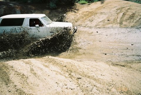 1990 Ford Bronco - white SUV throws up a heavy spray of mud as it powers across a rutted dirt and mud course.