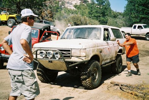 1990 Ford Bronco - Two men stand near a dusty white Ford Bronco fitted with a tube bumper and off-road lights at a tree-lined off-road staging area.