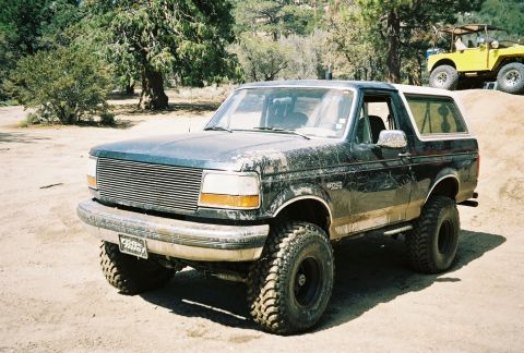 1990 Ford Bronco - lifted dark-green Ford Bronco with a billet grille and large mud-terrain tires sits on a dirt clearing with a yellow Jeep in the background.