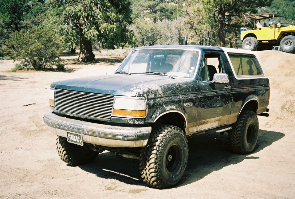 Lifted dark-green Ford Bronco with billet grille and mud-streaked body parked on dirt, yellow Jeep behind
