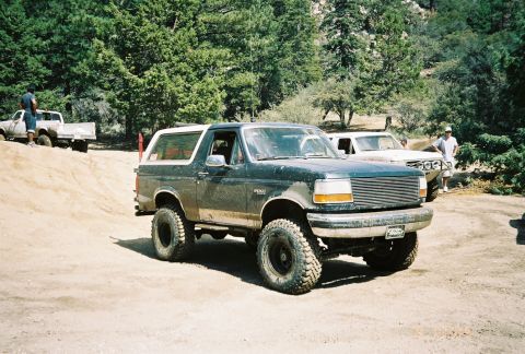 1990 Ford Bronco - lifted green Ford Bronco on oversized mud tires rests on a dirt mound in a forested off-road area alongside other parked vehicles.
