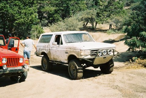 1990 Ford Bronco - mud-splattered white Ford Bronco with a tubular front bumper and off-road lights sits on a dirt trail next to a red Jeep and a man.