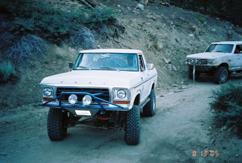 1990 Ford Bronco - white classic Ford pickup with a blue tube bumper and three off-road lights climbs a rocky mountain trail with another truck behind it.
