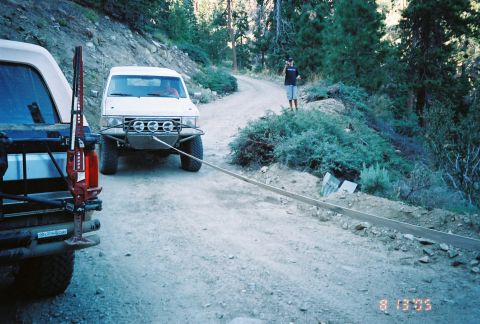 1990 Ford Bronco - white off-road truck is connected by a tow strap to another vehicle on a forested dirt trail while a person stands at the roadside.