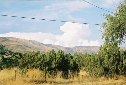 1990 Ford Bronco - Towering white cumulus clouds build over golden grassy hills lined with pine trees behind a wire fence and overhead wire.