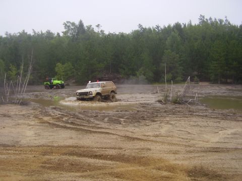 1995 Ford Bronco - A tan SUV navigates a muddy track, kicking up water, with a green off-road vehicle visible in the distance.