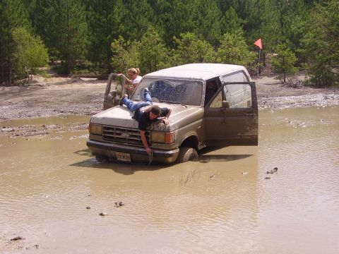 1995 Ford Bronco - A Ford Bronco is stuck in a mud pit with two people on the hood and in the driver's seat.