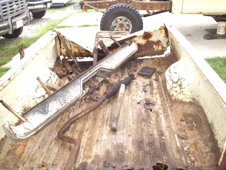 Rusty truck bed with bumper and exhaust parts. Another truck is partially visible
