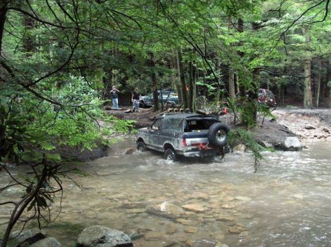 1995 Ford Bronco - Several off-road vehicles navigate a shallow stream in a wooded area.