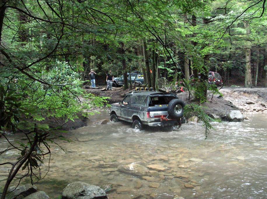 Ford Bronco driving through a stream with trees and other vehicles in the background