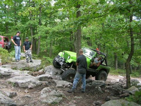 1995 Ford Bronco - Two Jeeps off-road on a rocky, wooded trail with several people watching.