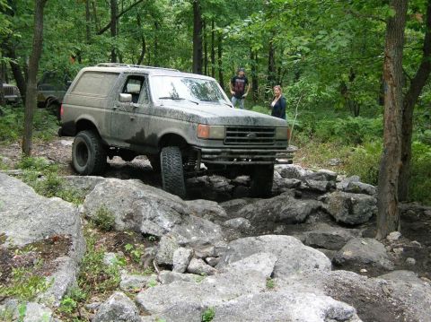 1995 Ford Bronco - A muddy silver-gray full-size Ford Bronco with a matching hardtop climbing over a rock ledge on a wooded trail; a man and a woman stand bes…