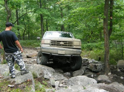 1995 Ford Bronco - A tan Ford Bronco is shown traversing a challenging rocky terrain in a forest.
