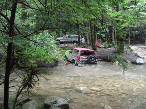 1995 Ford Bronco - A 1995 Ford Bronco navigates a rocky stream in a wooded area, with another vehicle following closely behind.