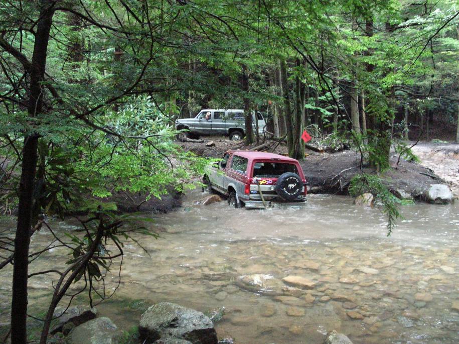 Red and silver 1995 Ford Bronco driving through a stream with another SUV behind it