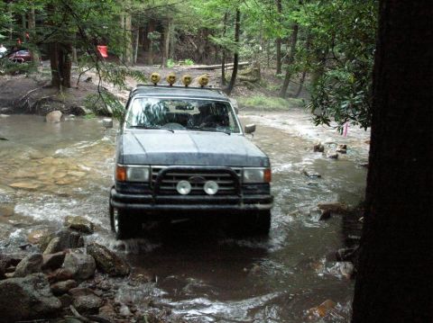 1995 Ford Bronco - Gray full-size Ford Bronco fording a shallow rocky creek in dense green woods, photographed head-on as water breaks around the front tires.…