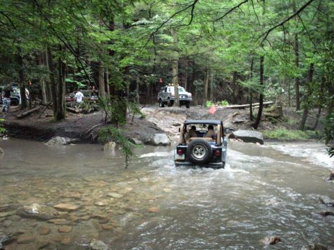 1995 Ford Bronco - Rear view of a topless Jeep Wrangler with tan interior and a tailgate-mounted spare fording a rocky, fast-running creek in the woods, water…