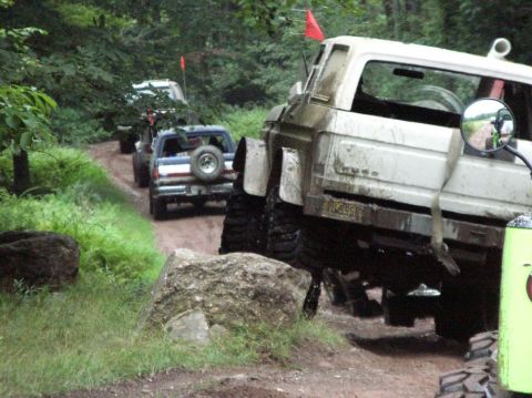 1995 Ford Bronco - Several off-road vehicles navigate a muddy trail surrounded by trees.