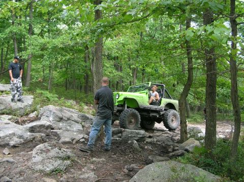 1995 Ford Bronco - A lime green open-top Jeep CJ on oversized mud tires and steel wheels crawls a rocky ledge on a wooded trail, fitted with a roll cage and n…