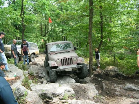 1995 Ford Bronco - A tan Jeep Wrangler is shown off-roading on a rocky, wooded path with other vehicles nearby.