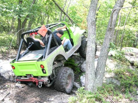 1995 Ford Bronco - Off-road vehicle with a roll cage navigates a challenging rocky and muddy trail through a wooded area.