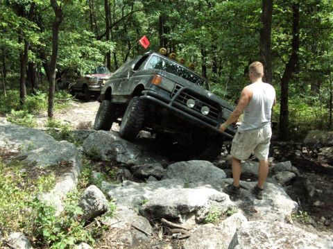 1995 Ford Bronco - A dark, muddy full-size Ford Bronco fitted with a front brush guard, roof-mounted round off-road lights and a tall red trail flag, tilting…