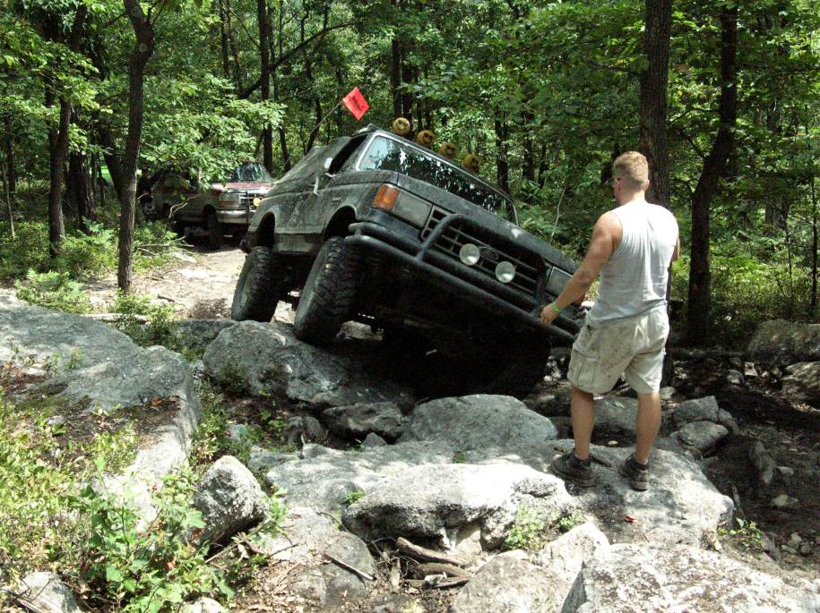 Dark full-size Ford Bronco with brush guard climbing rocks, a man spotting in front